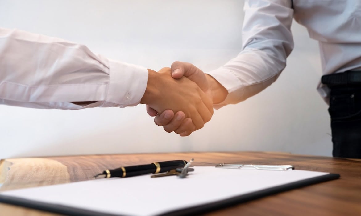 Zweibeiniger Händedruck bei Geschäftsabschluss auf Tisch, Dokumente und Stifte für Vertragsunterzeichnung sichtbar.