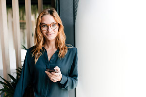 Inspirierte Frau mit Smartphone in formeller Kleidung vor Fenster, modernes Business-Umfeld.