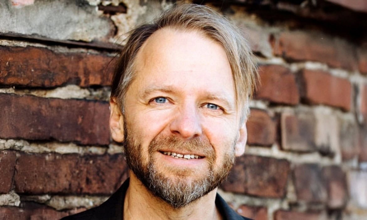 Einen professionellen Mann vor einer Ziegelsteinmauer, freundlich lächelnd, seriöse Ausstrahlung.