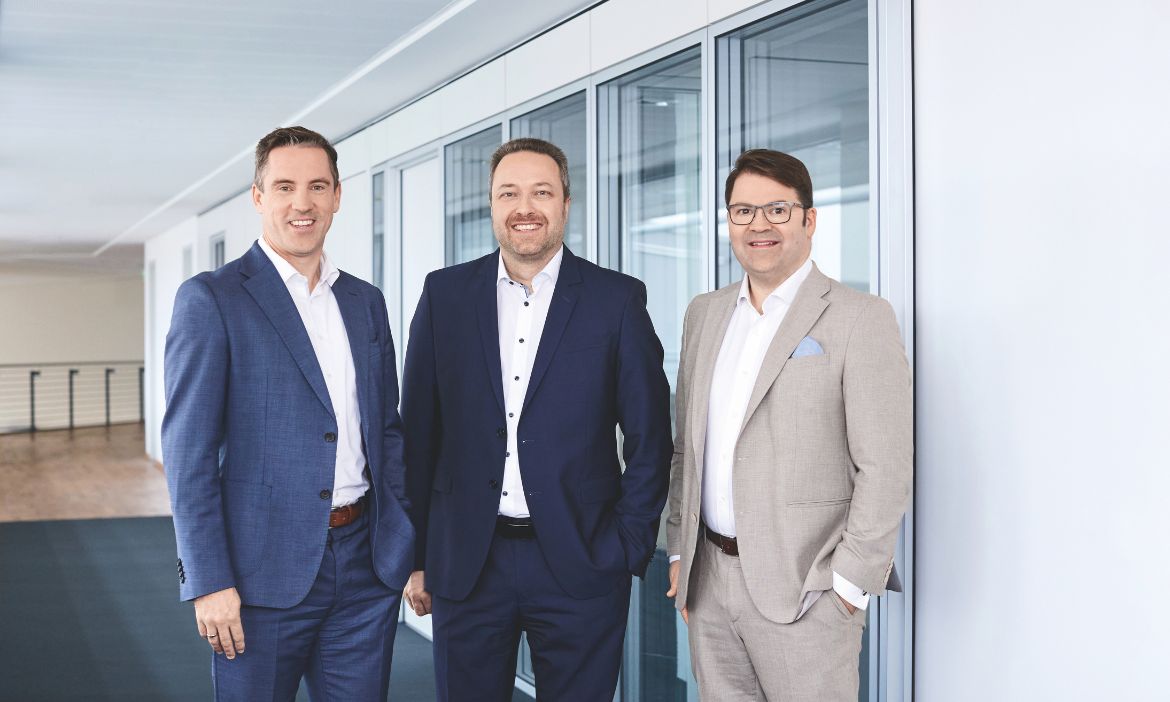 Männer in Business-Anzügen vor großen Fenstern im Bürogebäude.