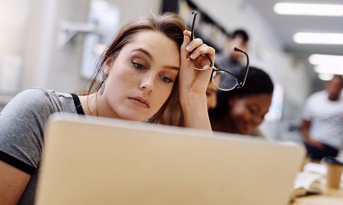 Junge Frau mit Brille, die gestresst am Schreibtisch sitzt, umgeben von anderen Schülern.