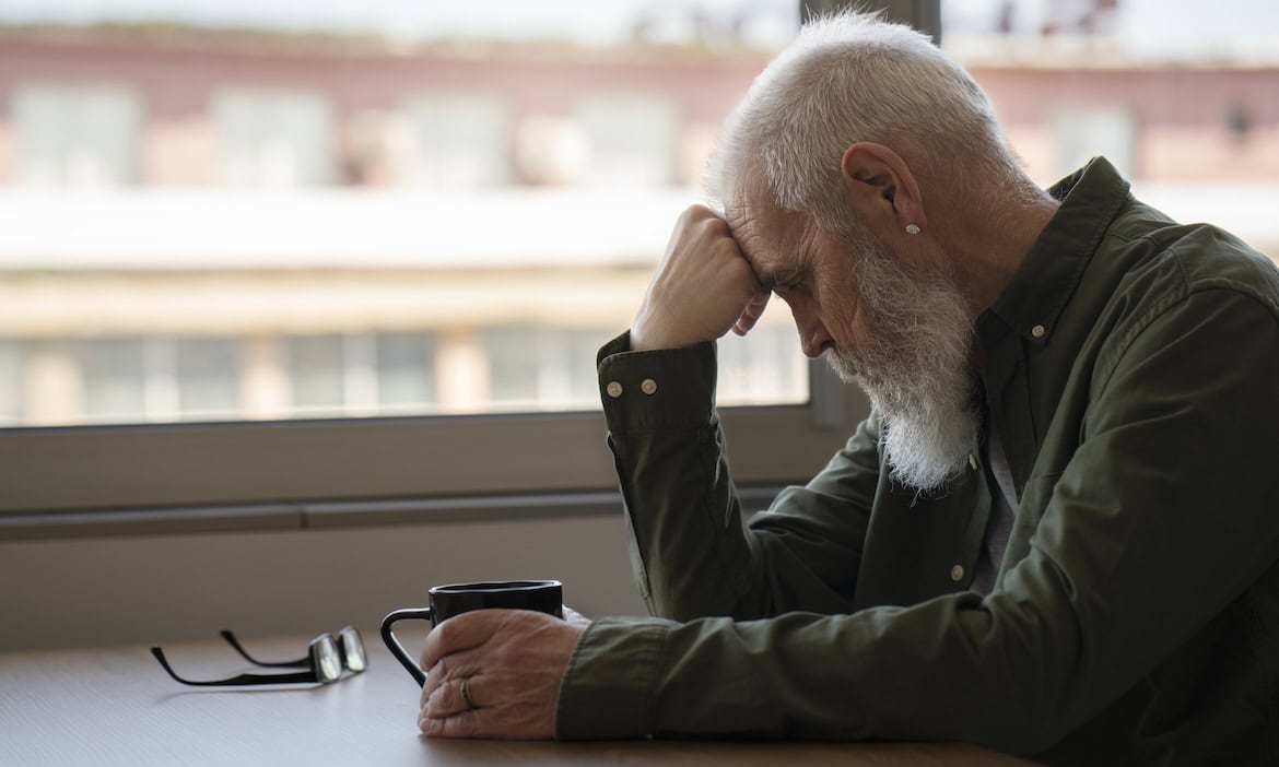 Älterer Mann mit Sorgen um seine Rente, sitzt nachdenklich am Fenster.